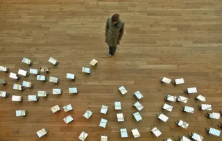 Vue de haut d'un parquet clair : une personne en manteau se tient debout au milieu, entour&eacute;e d'une cinquantaine de petits miroirs pos&eacute;s au sol et reli&eacute;s par des c&acirc;bles, tous orient&eacute;s vers elle.