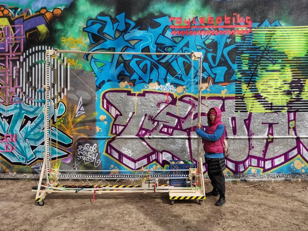 Un portique de bois mont&eacute; sur roulettes, dress&eacute; contre un mur couvert de graffitis ; une personne se tient &agrave; c&ocirc;t&eacute;, des bombes de peinture sont pos&eacute;es sur le rail horizontal