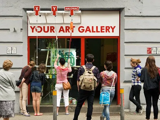 Une devanture peinte en rouge o&ugrave; on lit &laquo; Your Art Gallery &raquo; ; sur le trottoir, des passants tirent sur des cordes suspendues &eacute;tiquet&eacute;es X, Y et &laquo; malować &raquo;