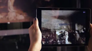 Dans la p&eacute;nombre de la salle, une main tient une tablette devant &laquo; L'Atelier du peintre &raquo; ; sur l'&eacute;cran, la toile est reprise et augment&eacute;e d'une &eacute;tiquette qui nomme un des personnages