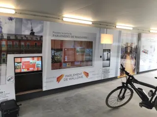 Une paroi de cloison imprim&eacute;e dans un couloir b&eacute;tonn&eacute;, sous des n&eacute;ons. Au centre, un grand &eacute;cran tactile paysage encastr&eacute; affiche une page d'accueil ; &agrave; droite, un &eacute;cran vertical plus &eacute;troit. Sur la paroi, en grandes lettres, &laquo; Poussez la porte du PARLEMENT DE WALLONIE ! &raquo;, et autour, en gris, les mots proposition, r&eacute;solution, d&eacute;cret, pr&eacute;sident, d&eacute;mocratie, auditions, contr&ocirc;le.