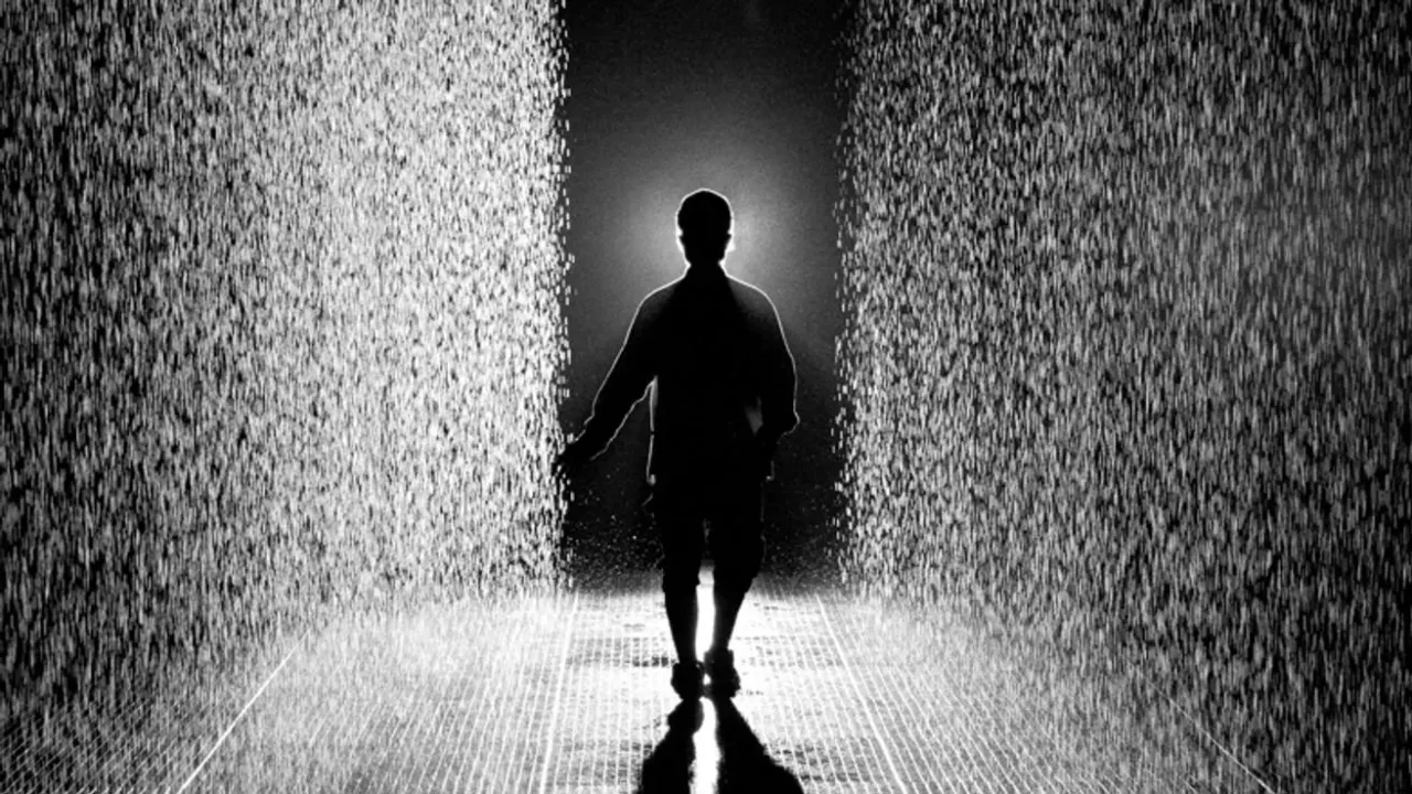 Photographie en noir et blanc : une silhouette vue de dos avance dans un couloir de pluie dense &eacute;clair&eacute; &agrave; contre-jour ; le sol sous ses pieds est sec.