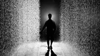 Photographie en noir et blanc : une silhouette vue de dos avance dans un couloir de pluie dense &eacute;clair&eacute; &agrave; contre-jour ; le sol sous ses pieds est sec.