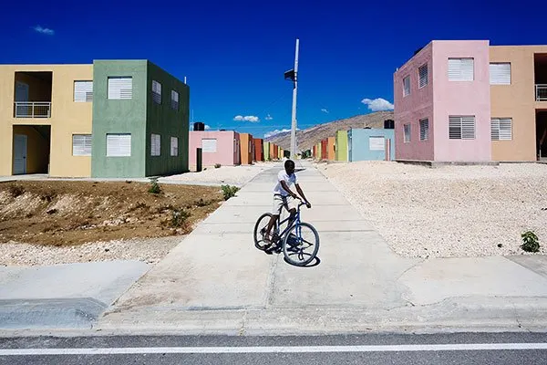 Un enfant &agrave; v&eacute;lo s'arr&ecirc;te au bord d'une rue neuve bord&eacute;e de petits immeubles peints en vert, rose et ocre, sous un ciel bleu