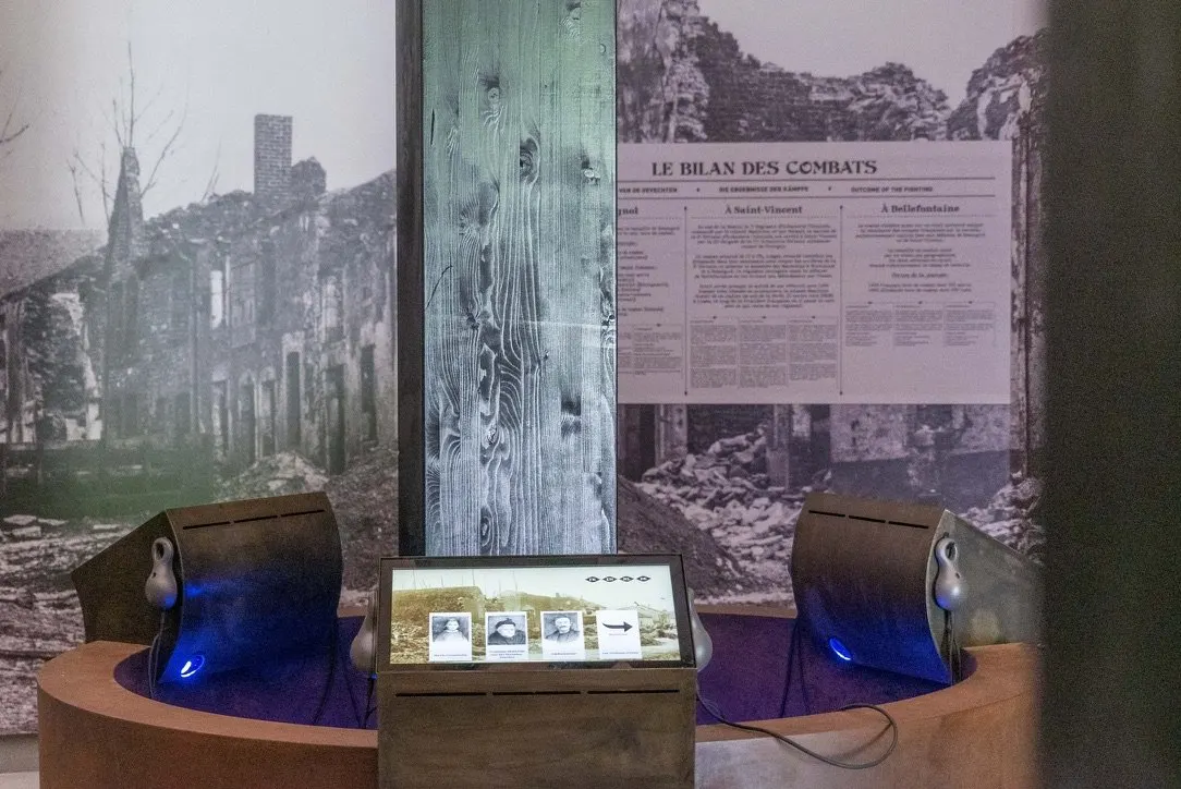 Deux pupitres de m&eacute;tal rouill&eacute; se faisant face de part et d'autre d'un poteau de bois clair, chacun avec un combin&eacute; d'&eacute;coute gris suspendu et une lumi&egrave;re bleue &agrave; sa base. Entre eux, un troisi&egrave;me &eacute;cran sur socle de bois affiche une photographie ancienne de tranch&eacute;e et une rang&eacute;e de portraits. Derri&egrave;re, une paroi imprim&eacute;e montre des ruines de village et un panneau intitul&eacute; &laquo; LE BILAN DES COMBATS &raquo;.