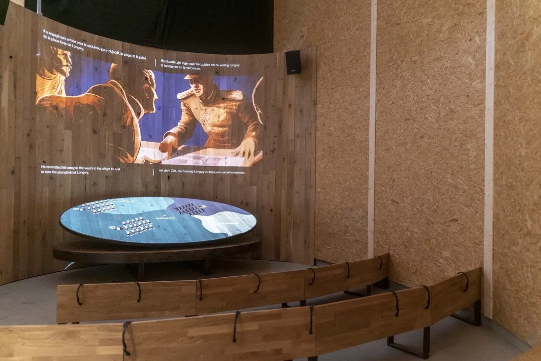 Une salle de projection aux parois de bois clair. Sur un mur courbe, une animation en tons ocre et bleus montre deux officiers pench&eacute;s sur une carte ; autour de l'image, quatre blocs de sous-titres en fran&ccedil;ais, n&eacute;erlandais, anglais et allemand. Devant l'&eacute;cran, une table ovale affiche une carte lumineuse bleue sem&eacute;e de petits marqueurs de troupes. Deux rang&eacute;es de bancs de bois incurv&eacute;s font face &agrave; l'ensemble.