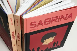 Trois exemplaires du livre pos&eacute;s debout, l'un ouvert en &eacute;ventail ; la couverture rose et rouge montre un visage de profil au-dessus d'un rang de silhouettes noires.