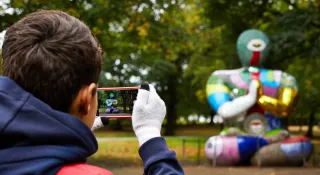 Un visiteur de dos l&egrave;ve son t&eacute;l&eacute;phone vers une sculpture color&eacute;e pos&eacute;e dans l'herbe du parc ; l'&eacute;cran affiche la silhouette &agrave; faire co&iuml;ncider avec elle