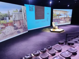 Salle de spectacle sombre : trois grands &eacute;crans en arc de cercle, l'un montrant un jardin avec du linge qui s&egrave;che, l'autre l'interface bleue de GaIA &mdash; une grille de cercles et des relev&eacute;s techniques &mdash;, le troisi&egrave;me une terrasse en bord de mer. Devant, une estrade en bois &eacute;clair&eacute;e avec un fauteuil blanc ; au premier plan, des rang&eacute;es de si&egrave;ges en plastique moul&eacute;.