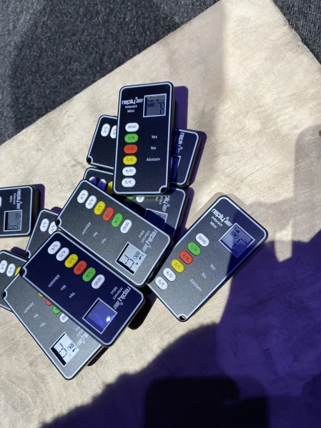 Une dizaine de t&eacute;l&eacute;commandes grises et noires empil&eacute;es sur une planche de bois : petit &eacute;cran &agrave; cristaux liquides, un bouton &laquo; send &raquo; et cinq touches color&eacute;es num&eacute;rot&eacute;es 1/A &agrave; 5/E, avec les mentions &laquo; Yes &raquo;, &laquo; No &raquo; et &laquo; Abstain &raquo; s&eacute;rigraphi&eacute;es &agrave; c&ocirc;t&eacute;. La marque &laquo; reply Interact Mini &raquo; figure en haut.