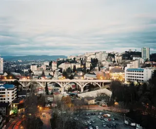 Vue de Sotchi au cr&eacute;puscule : un viaduc &agrave; arches blanches traverse un vallon, des immeubles blancs et des toits de t&ocirc;le escaladent la colline jusqu'aux montagnes, un parking &eacute;clair&eacute; au premier plan.