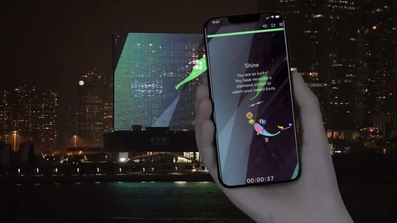 Une main tient un t&eacute;l&eacute;phone o&ugrave; nage un poisson dessin&eacute; ; derri&egrave;re, de l'autre c&ocirc;t&eacute; du port de Hong Kong, la fa&ccedil;ade LED du mus&eacute;e M+ affiche le m&ecirc;me &eacute;tang la nuit