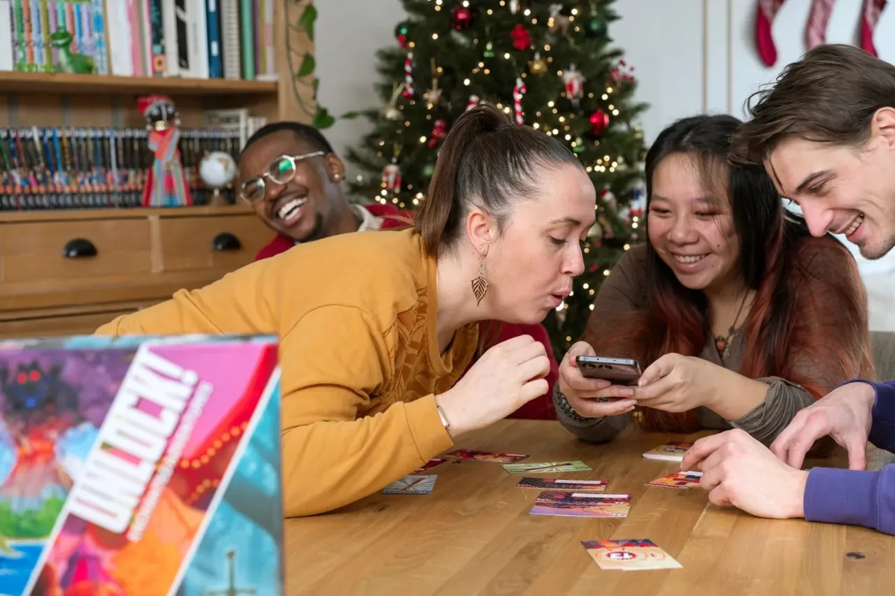 Quatre personnes pench&eacute;es autour d'une table de salon couverte de cartes &eacute;tal&eacute;es ; l'une d'elles tient un t&eacute;l&eacute;phone que les autres regardent