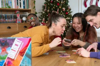 Quatre personnes pench&eacute;es autour d'une table de salon couverte de cartes &eacute;tal&eacute;es ; l'une d'elles tient un t&eacute;l&eacute;phone que les autres regardent
