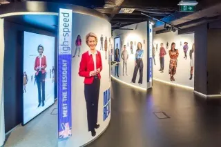 Couloir courbe d'un centre de visite, aux parois blanches. &Agrave; gauche, un grand &eacute;cran vertical montre une femme en veste rouge, debout, en pied. &Agrave; c&ocirc;t&eacute;, un bandeau bleu vertical porte les mots &laquo; MEET THE PRESIDENT &raquo;. Sur la paroi de droite, des silhouettes de personnes d&eacute;coup&eacute;es grandeur nature sont coll&eacute;es entre d'autres &eacute;crans verticaux. Un liser&eacute; lumineux court au pied des cloisons.