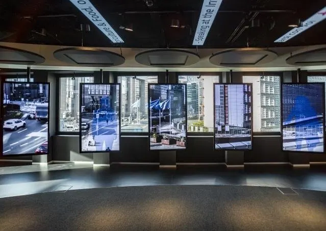 Une rang&eacute;e de six &eacute;crans verticaux align&eacute;s devant les baies vitr&eacute;es d'une salle sombre au plafond noir. Chacun montre une vue de rue du quartier europ&eacute;en : carrefours, immeubles de bureaux vitr&eacute;s, drapeaux bleus align&eacute;s. Au plafond, des panneaux acoustiques ovales et des bandeaux de texte imprim&eacute;s en diagonale.
