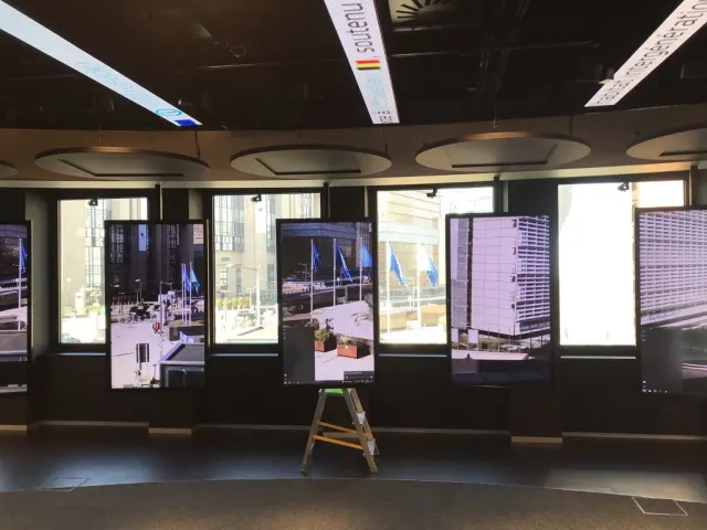 La m&ecirc;me salle aux &eacute;crans verticaux, photographi&eacute;e pendant l'installation : les moniteurs sont allum&eacute;s sur des images de rue, mais la pi&egrave;ce est sombre et un escabeau en bois est rest&eacute; plant&eacute; devant l'un d'eux, au milieu du passage.