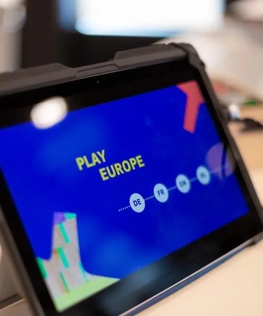 Gros plan sur une tablette durcie dans une coque noire, pos&eacute;e sur un plan de travail. L'&eacute;cran affiche sur fond bleu vif le titre &laquo; PLAY EUROPE &raquo; en lettres jaunes, et sous lui une rang&eacute;e de quatre pastilles rondes reli&eacute;es par des pointill&eacute;s : DE, FR, EN, NL.