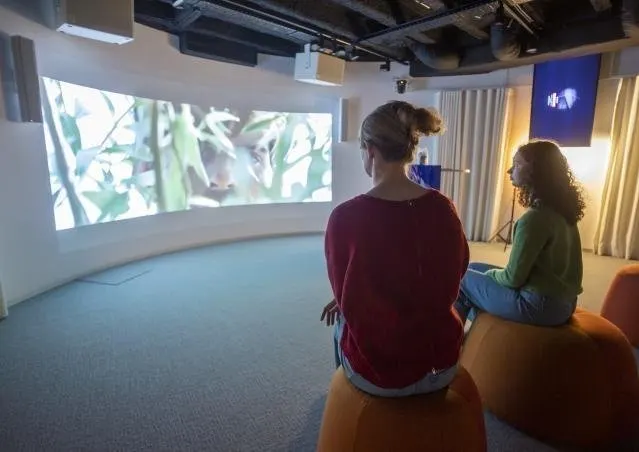 Deux femmes assises de dos sur des poufs orange dans une petite salle sombre, face &agrave; un &eacute;cran courbe qui occupe tout le mur et projette une image de feuillage vert. &Agrave; droite, un moniteur bleu plus petit et un rideau clair ; le sol est couvert d'une moquette grise.
