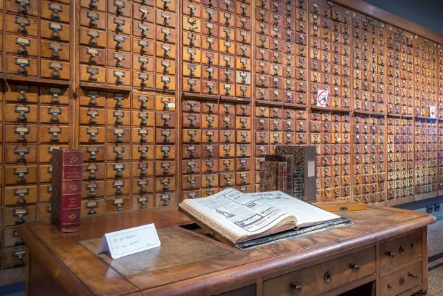 Un mur entier de tiroirs &agrave; fiches en bois clair, sur une dizaine de rang&eacute;es, chaque tiroir muni d'une poign&eacute;e m&eacute;tallique et d'un porte-&eacute;tiquette ; au premier plan un bureau ancien avec un gros volume reli&eacute; ouvert sur un lutrin, deux registres debout et un carton manuscrit &laquo; Ne pas toucher ! Ne rien d&eacute;poser ! &raquo;.